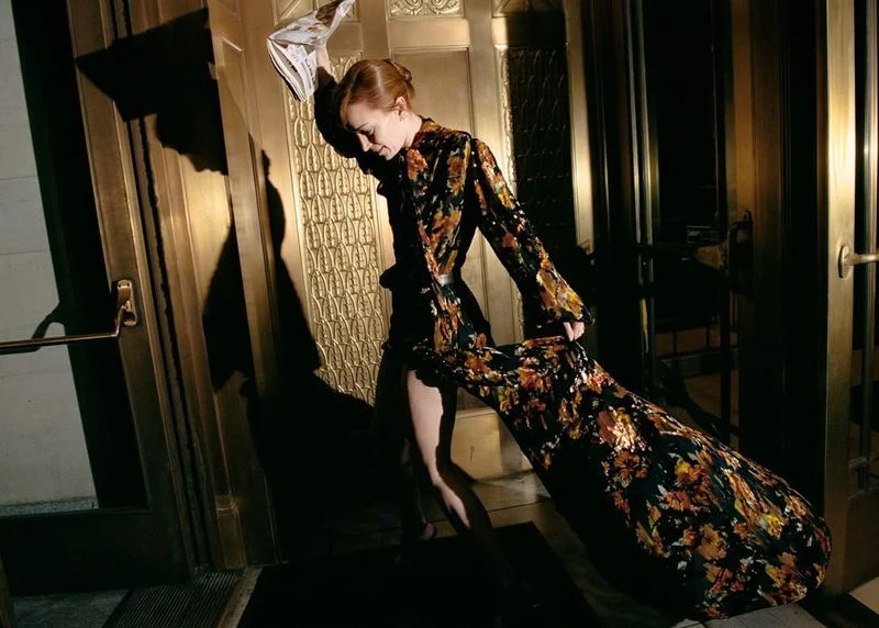 Elegant woman in floral gown stands in a doorway with gilded doors, holding a newspaper, creating a dramatic, fashionable composition.