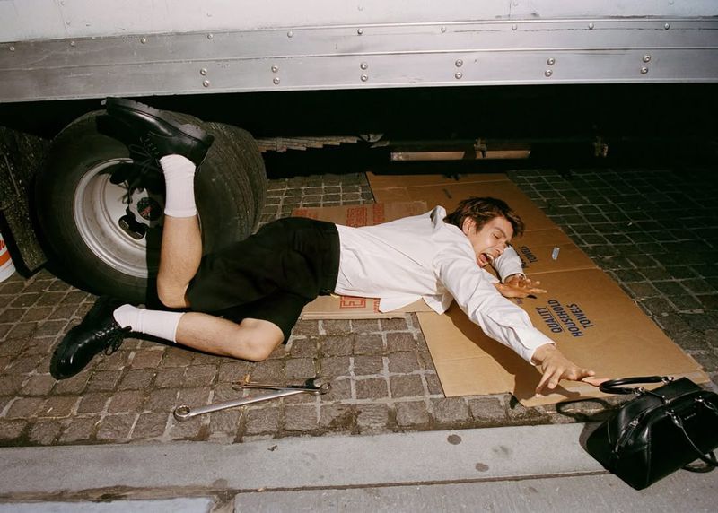 A man lying on cardboard under a truck near its tire with hand tools on cobblestone, wearing a white button-down shirt and dark shorts.