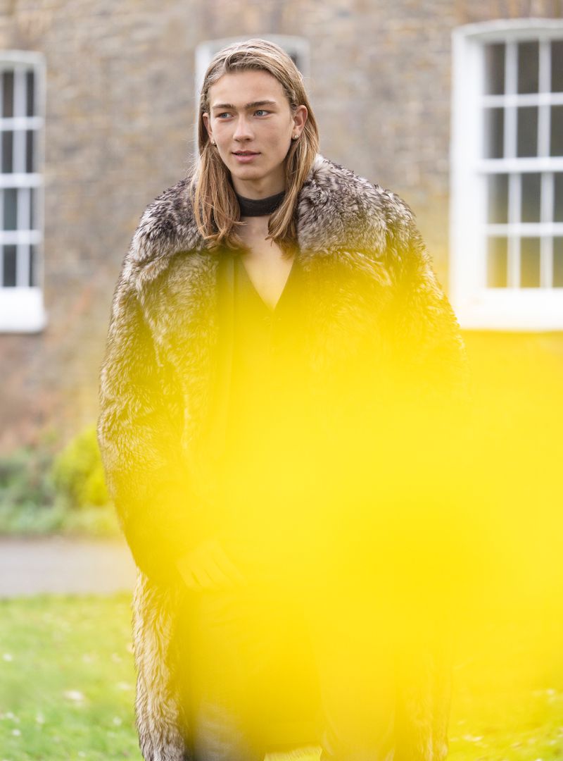 Model with long blond hair wearing a fur coat outdoors against a stone building with white windows