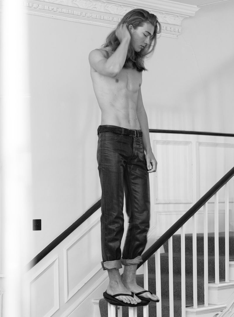 Shirtless male model with long hair standing on a staircase wearing dark trousers in a black and white editorial photograph.