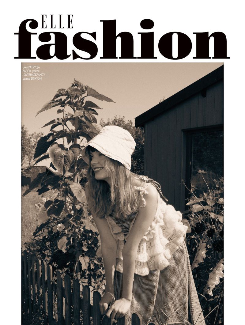 Monochrome fashion editorial image featuring a smiling woman in a bucket hat leaning on a wooden fence near sunflowers and a rustic building.