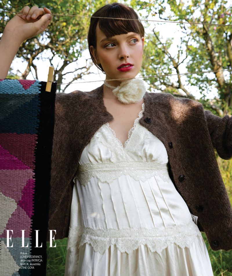 Model in white lace dress and brown cardigan standing outdoors near laundry line with colorful textile