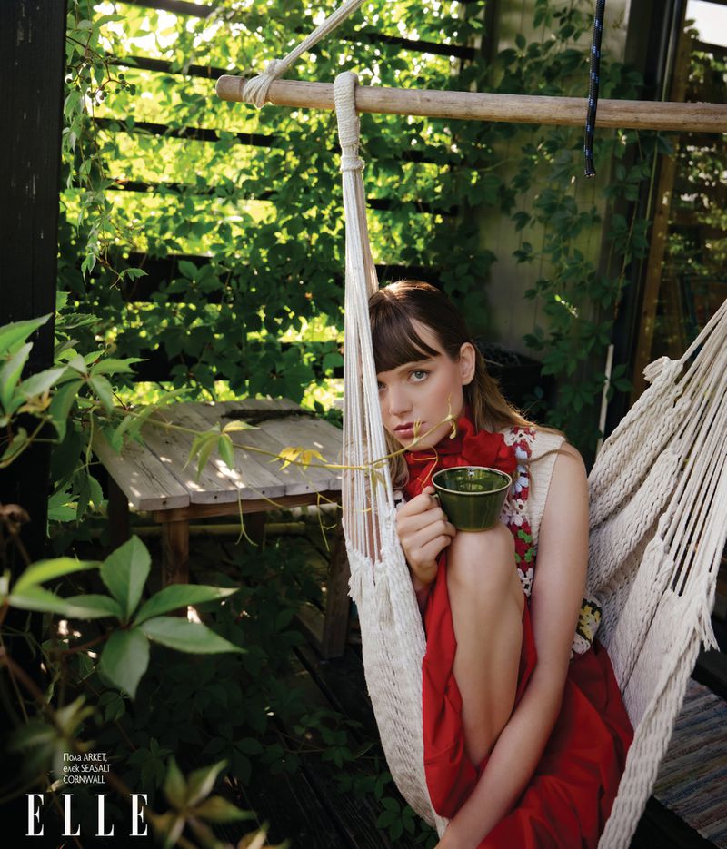 Woman sitting in a white woven hammock outdoors holding a green ceramic cup, surrounded by leafy plants and wooden fencing.