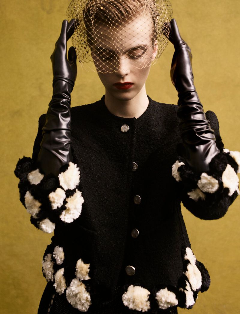 Model in black jacket with white pom poms and long leather gloves holding a veil over her face