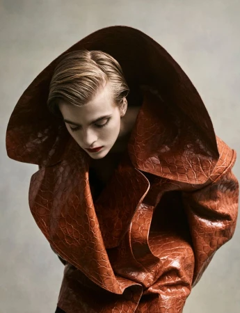 Model wearing a structured brown textured coat with an oversized exaggerated hood against a neutral studio backdrop