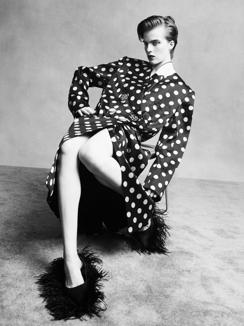 Model seated on a chair wearing a black and white polka dot coat and feathered shoes against a neutral studio backdrop.