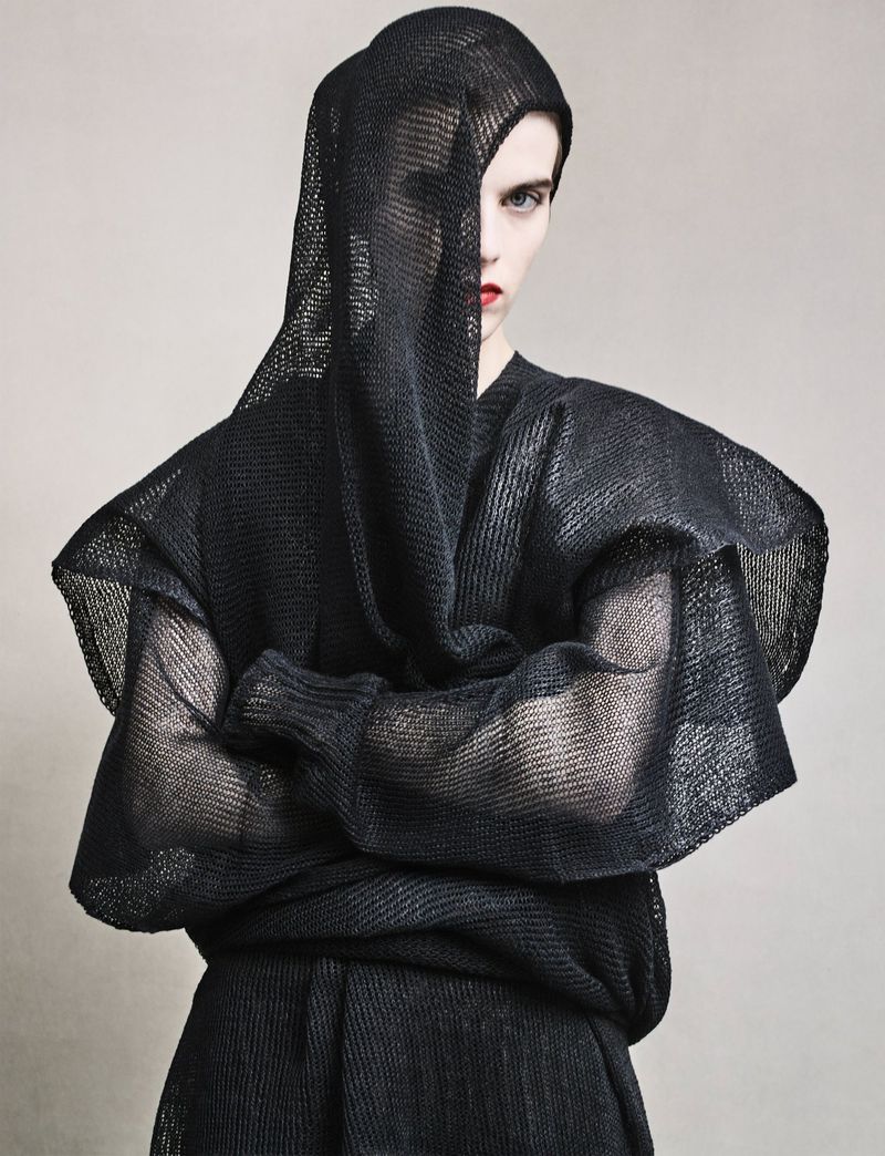 Model wearing a black mesh draped top with arms crossed against a neutral background