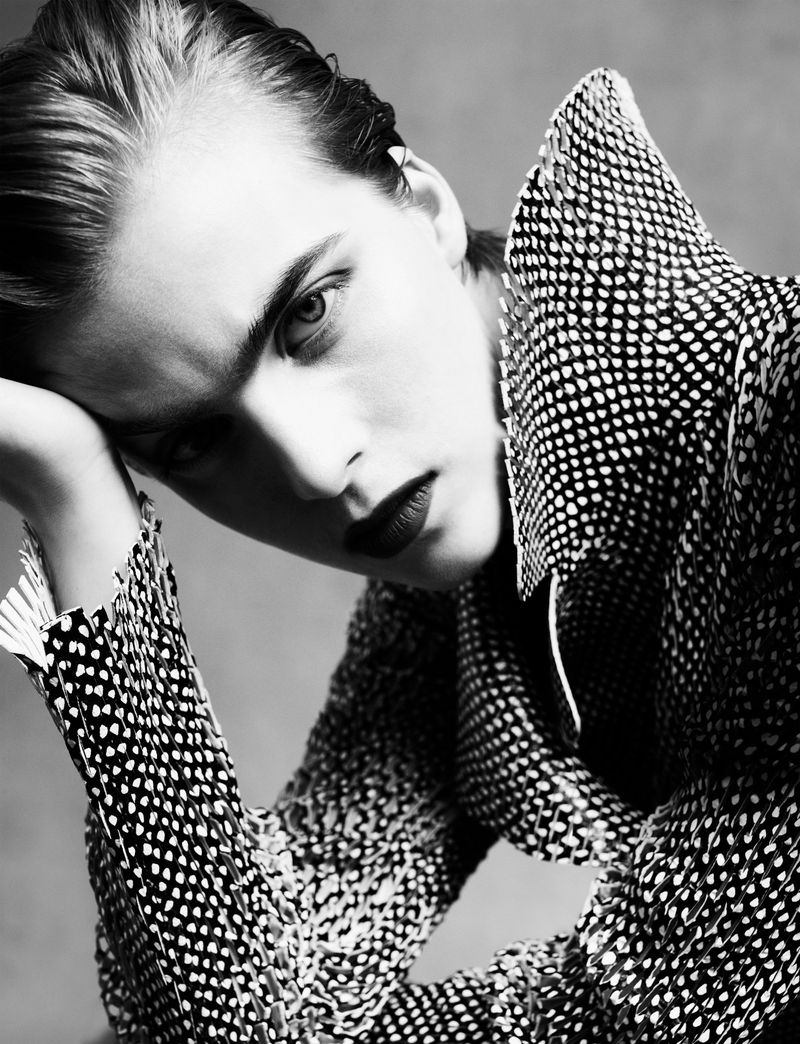 Close-up monochrome portrait of a model with dark lipstick wearing a patterned textured high-collar jacket.