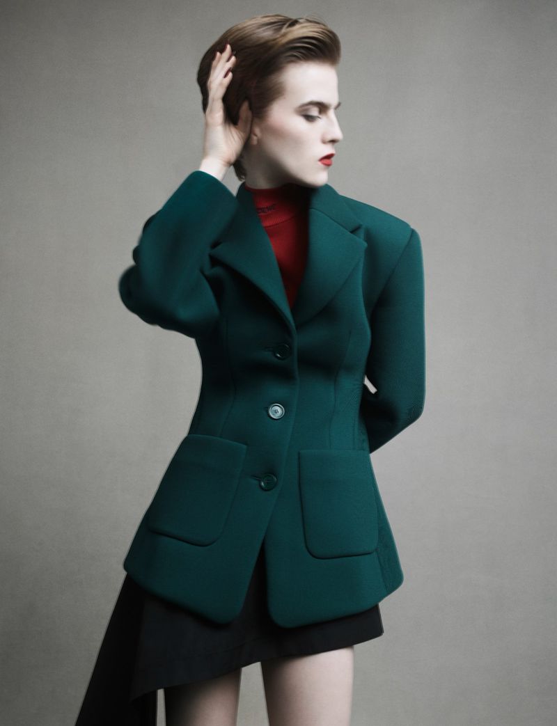Model with short hair wearing a structured green blazer and red top against a grey seamless background.