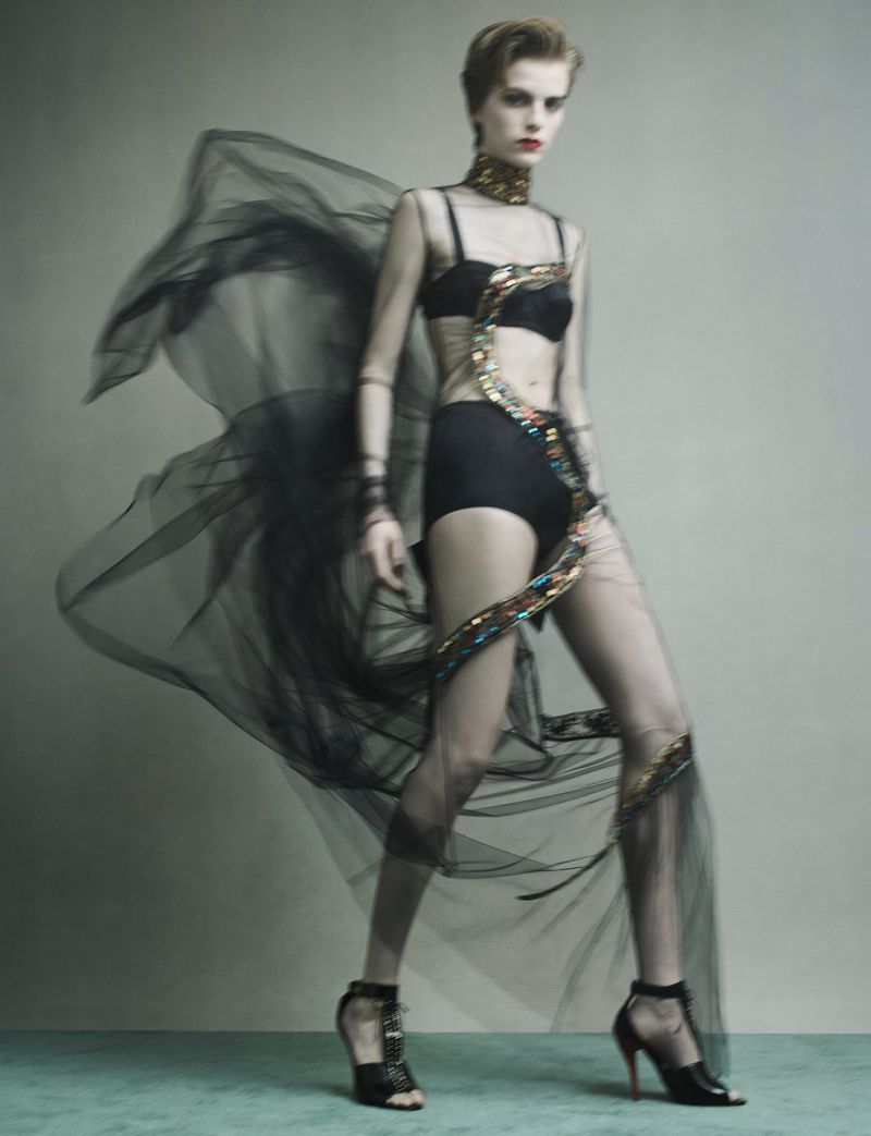 Fashion shoot of a slender woman in black lingerie and a sheer black cape, posing in a studio with a neutral background and dramatic lighting.
