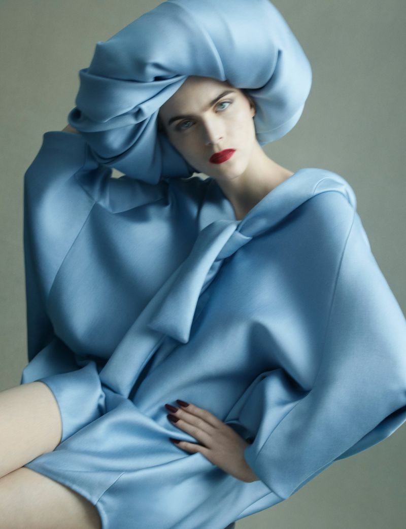 Model wearing a structured blue silk dress and oversized matching headpiece against a plain studio background.
