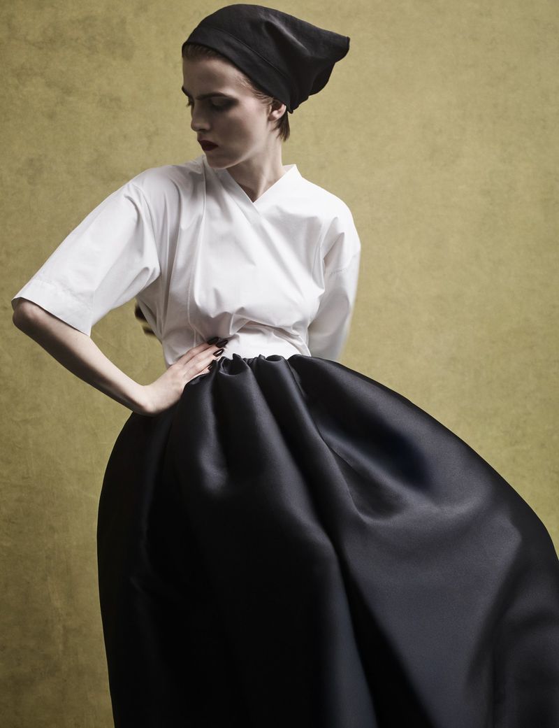 Model wearing a black headscarf, white top, and voluminous black skirt against a textured olive backdrop.