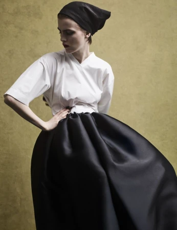 Model wearing a black headscarf, white top, and voluminous black skirt against a textured olive backdrop.