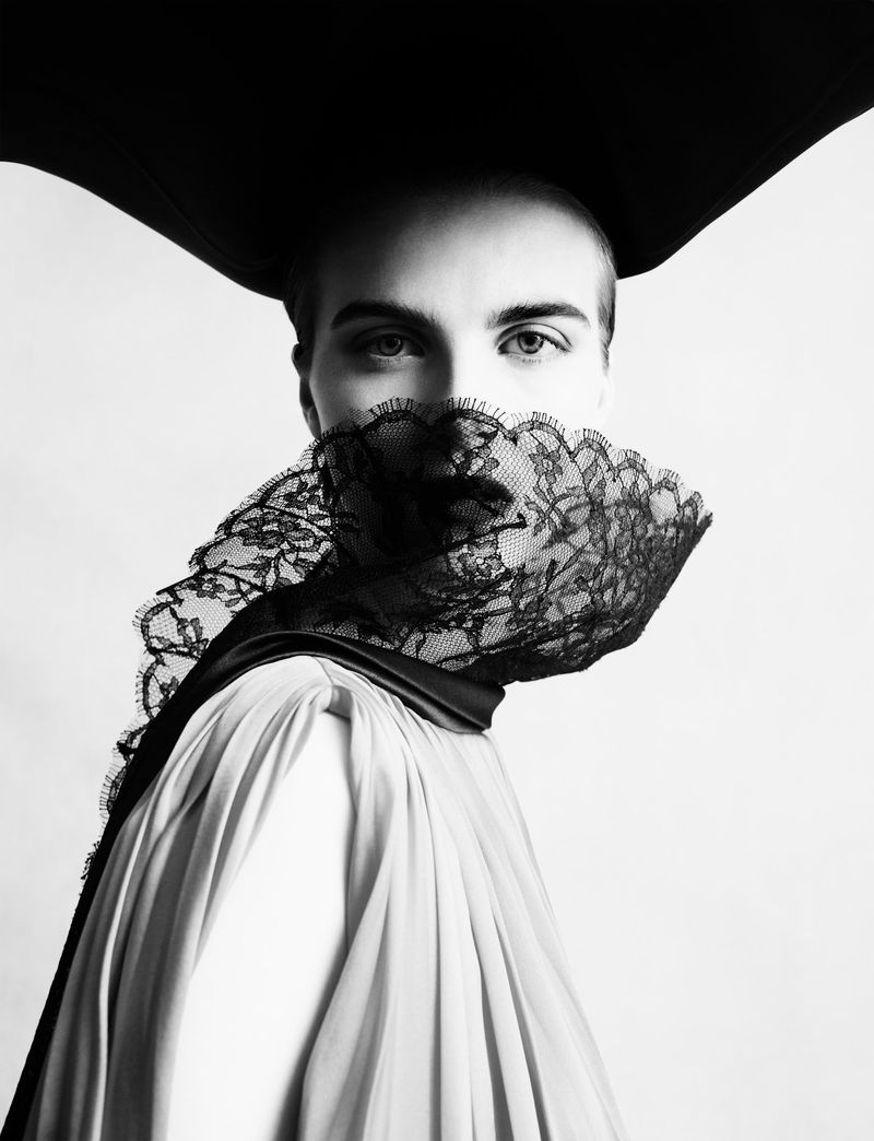 Model wearing a black lace face covering and large hat with a white garment against a seamless white studio background