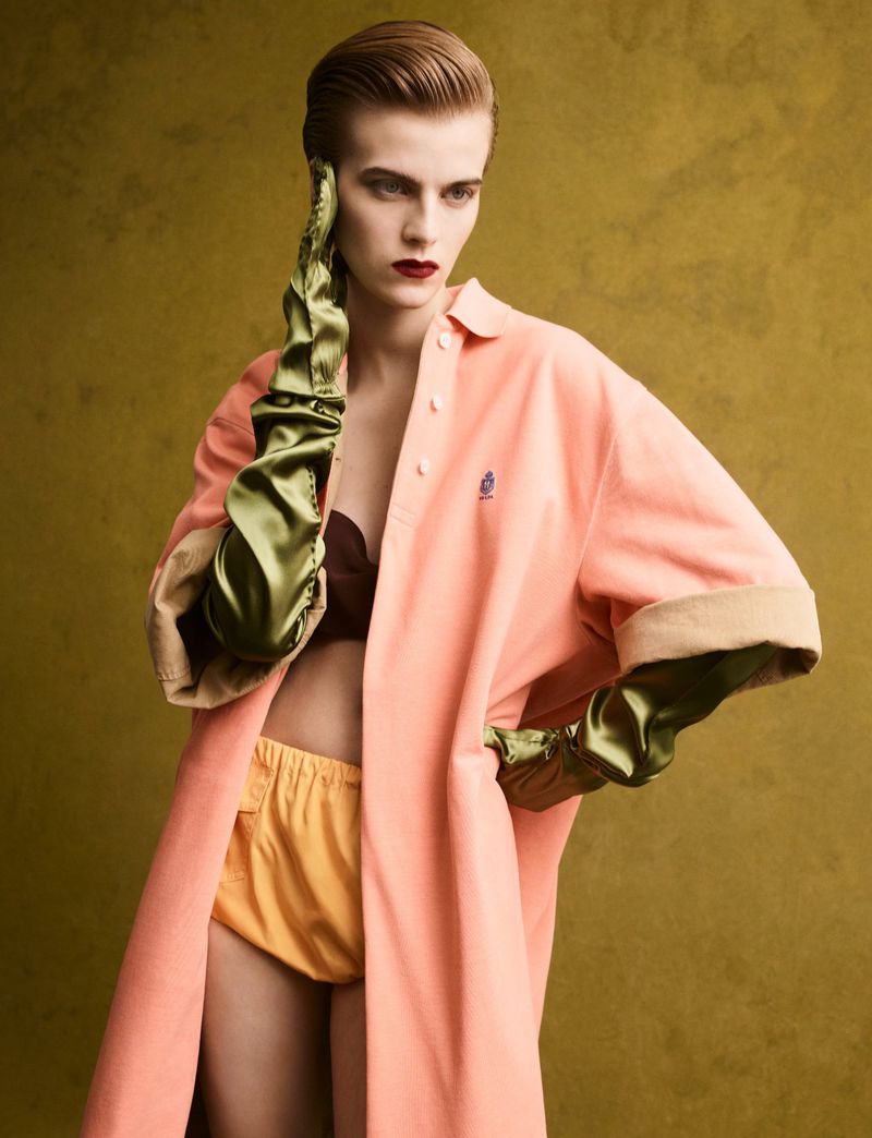 Model with slicked-back hair wearing a peach coat, yellow shorts, and long green satin gloves against a dark textured olive background