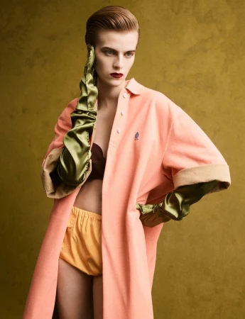 Model with slicked-back hair wearing a peach coat, yellow shorts, and long green satin gloves against a dark textured olive background