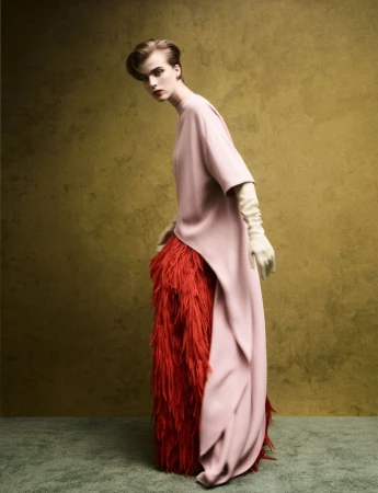 Model wearing a light pink tunic over a textured red feather skirt against a mottled olive green background.
