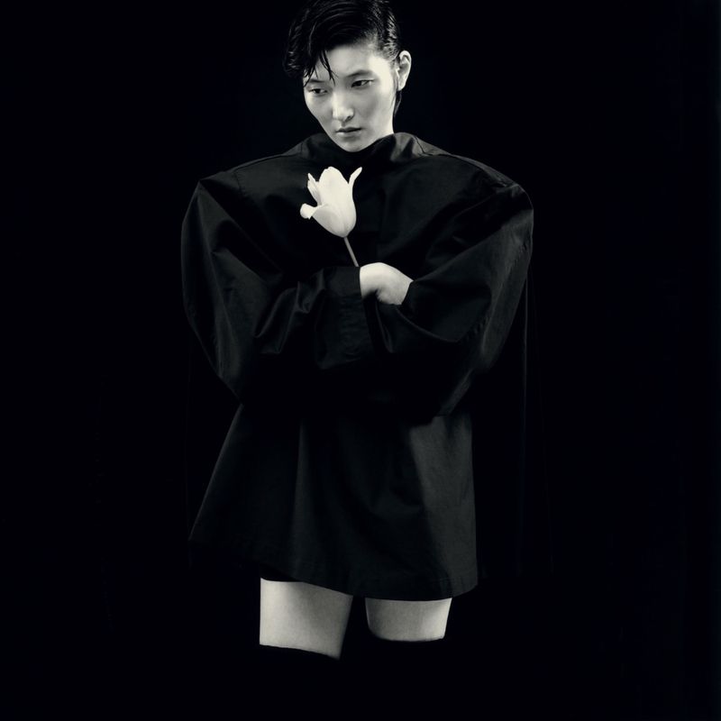 Model in black sculptural top holding white flower against dark background for Re-Edition Magazine