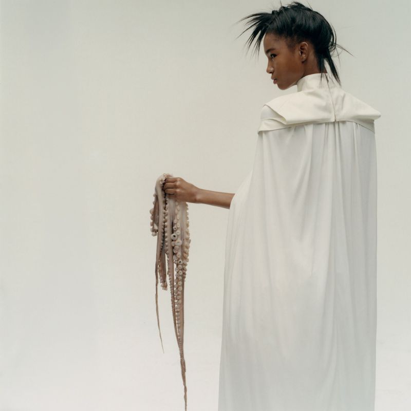Model in long white cape holding an octopus against a plain white studio background