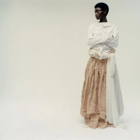 Model with short dark hair wearing a white long-sleeve top and layered tan skirt against a white studio background.