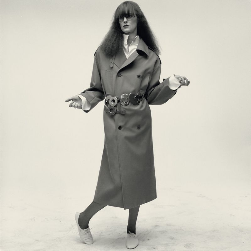 Model in a long trench coat with a statement belt and white sneakers posing against a white seamless background