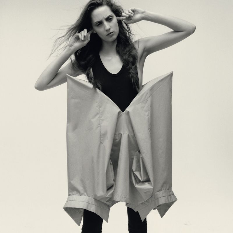 Model wearing a black top and structured sculptural garment against a light seamless background