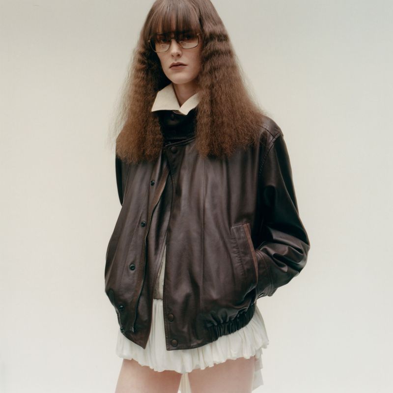 Model in tinted glasses wearing an oversized brown leather jacket over a white ruffled skirt against a plain backdrop.