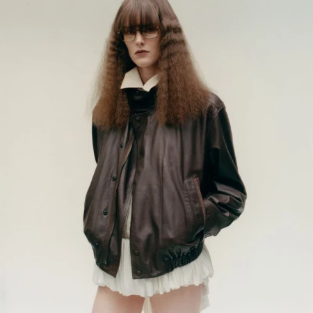 Model in tinted glasses wearing an oversized brown leather jacket over a white ruffled skirt against a plain backdrop.