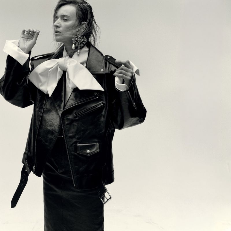 Model in black leather jacket with large white bow collar and statement earrings against a plain light background