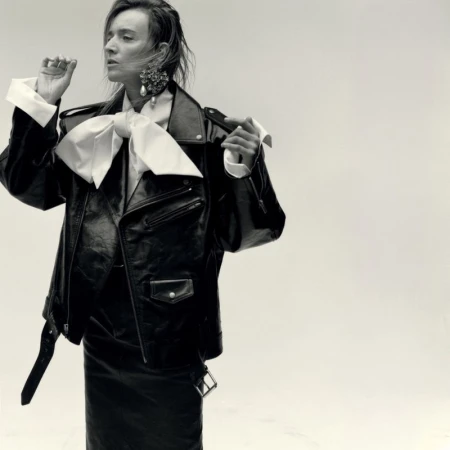 Model in black leather jacket with large white bow collar and statement earrings against a plain light background