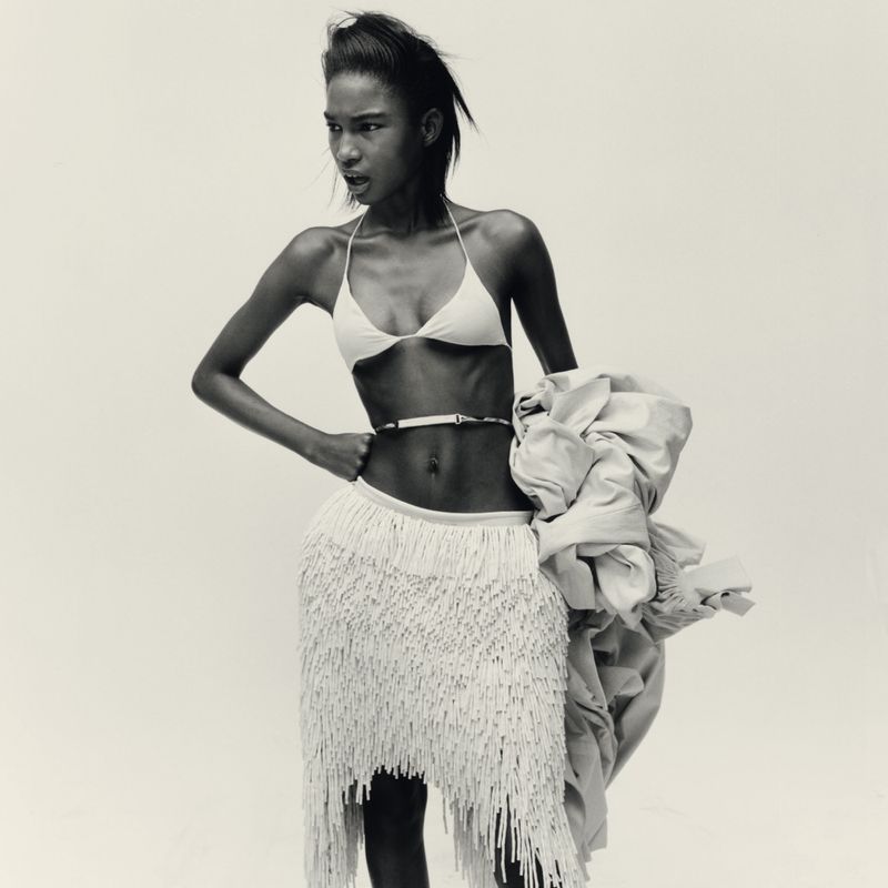An adult female model with dark hair poses in a fringed skirt and bikini top against a neutral backdrop in a fashion editorial shot.