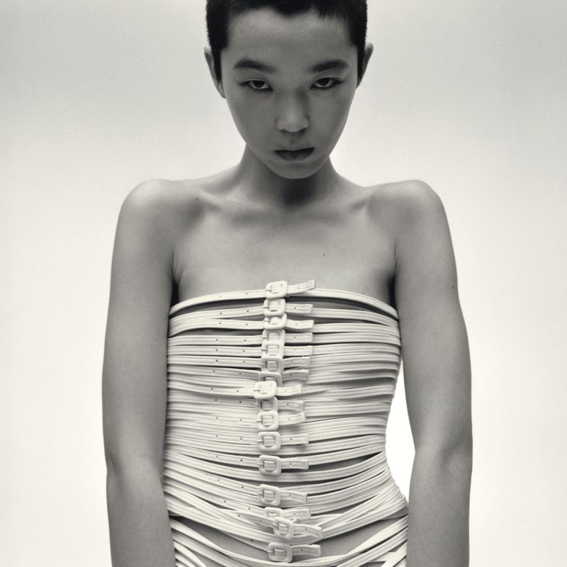 Model with short dark hair wearing a strapless top made of multiple buckled straps against a plain white background