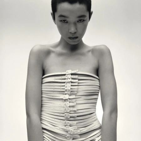 Model with short dark hair wearing a strapless top made of multiple buckled straps against a plain white background
