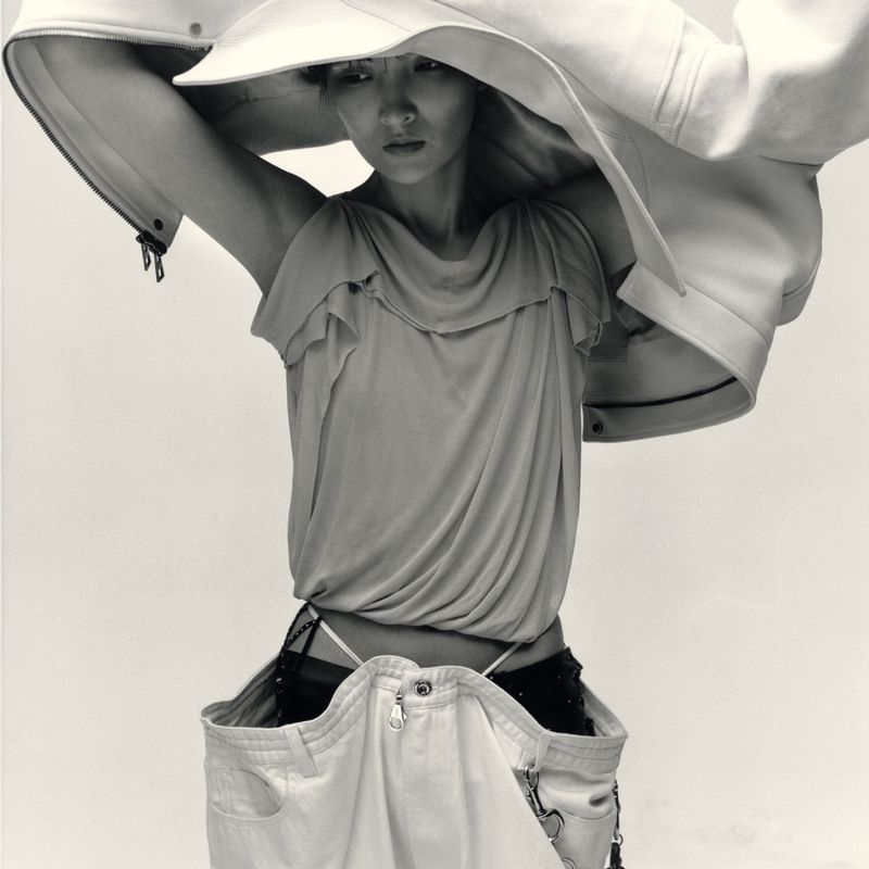 Monochrome fashion editorial portrait of a model lifting a structured white jacket overhead against a plain background