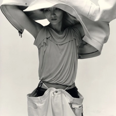 Monochrome fashion editorial portrait of a model lifting a structured white jacket overhead against a plain background