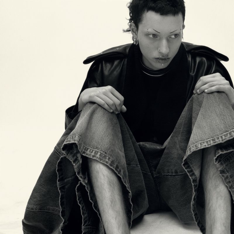Model wearing dark jacket and wide denim shorts seated on a plain white background in black and white