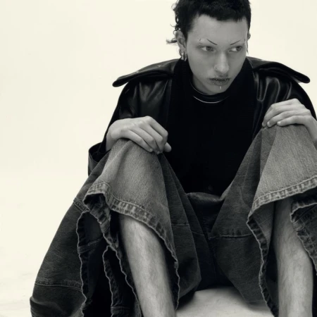 Model wearing dark jacket and wide denim shorts seated on a plain white background in black and white