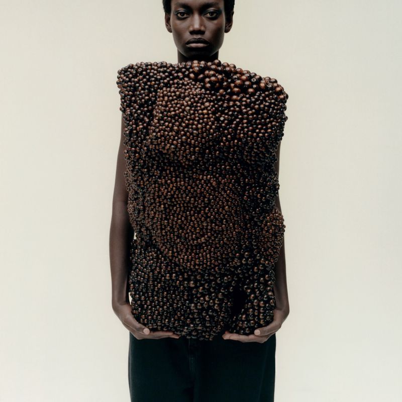 Model wearing a structured garment made entirely of small brown beads against a neutral white studio backdrop