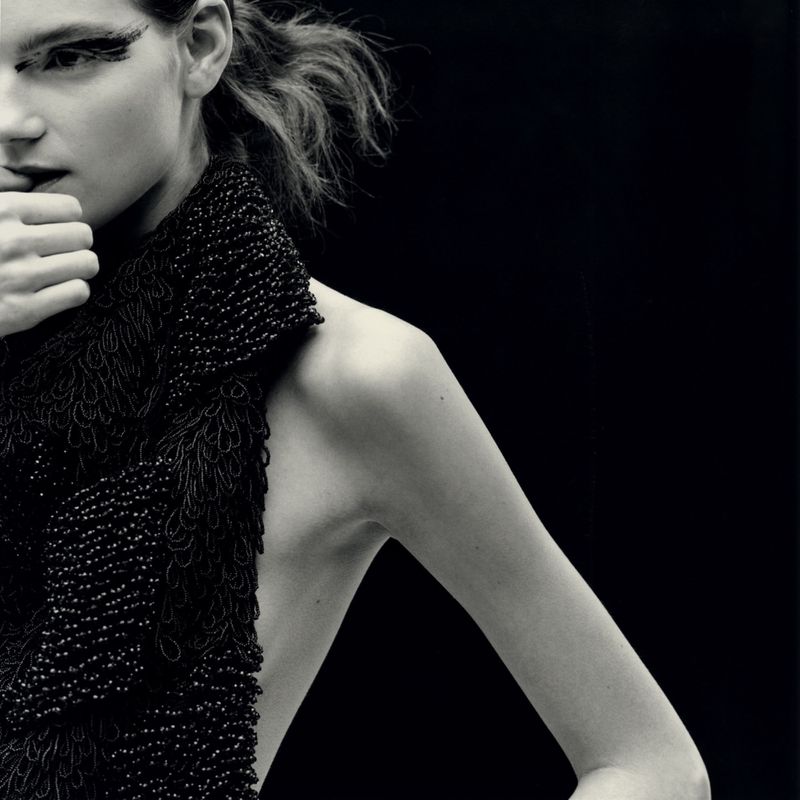 Close-up portrait of a model with dramatic eye makeup wearing a textured black beaded top against a dark background