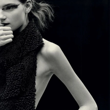 Close-up portrait of a model with dramatic eye makeup wearing a textured black beaded top against a dark background