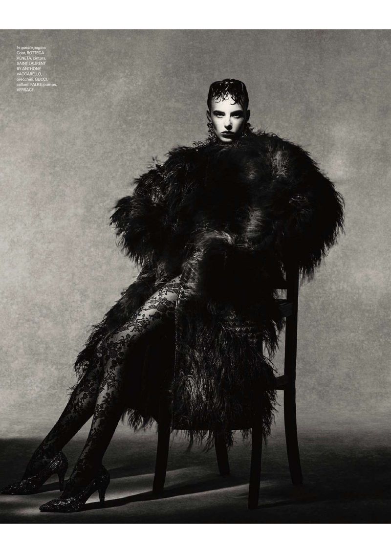 Model seated on a chair wearing a textured dark coat and patterned tights against a neutral textured studio background.