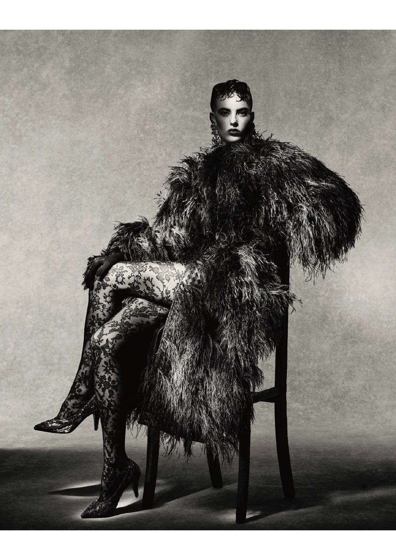 Model seated on a chair wearing a textured feathered coat and patterned tights against a neutral studio backdrop