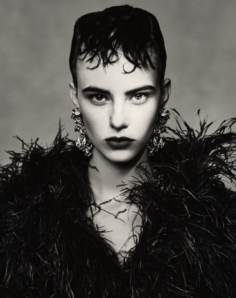 Monochrome portrait of a young woman with short hair, wearing large earrings and a feathered top against a grey backdrop.