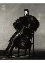Model seated on a chair wearing a textured dark coat and patterned tights against a neutral textured studio background.