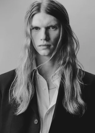 Portrait of a male model with long hair wearing an open white shirt and a dark blazer against a plain studio background.