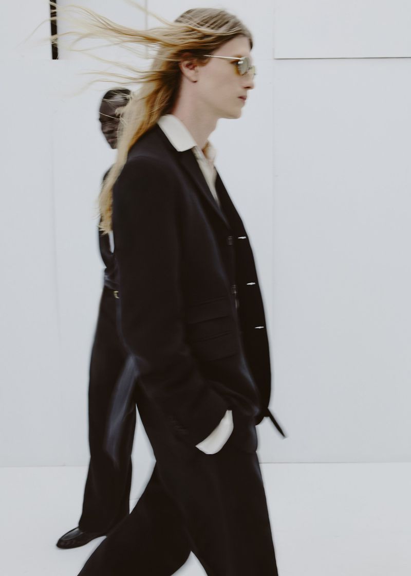 Long-haired male model wearing a dark suit jacket and sunglasses against a plain white background, with motion blur on his hair.
