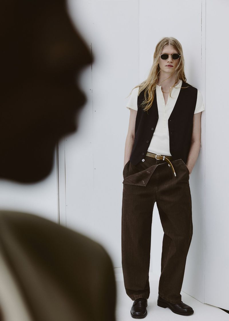 Model with long blonde hair wearing sunglasses, a dark vest, and trousers against a white backdrop