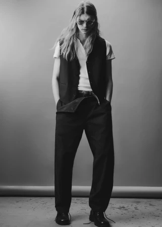 Model with long hair wearing a white t-shirt, dark vest, and trousers against a grey studio backdrop.