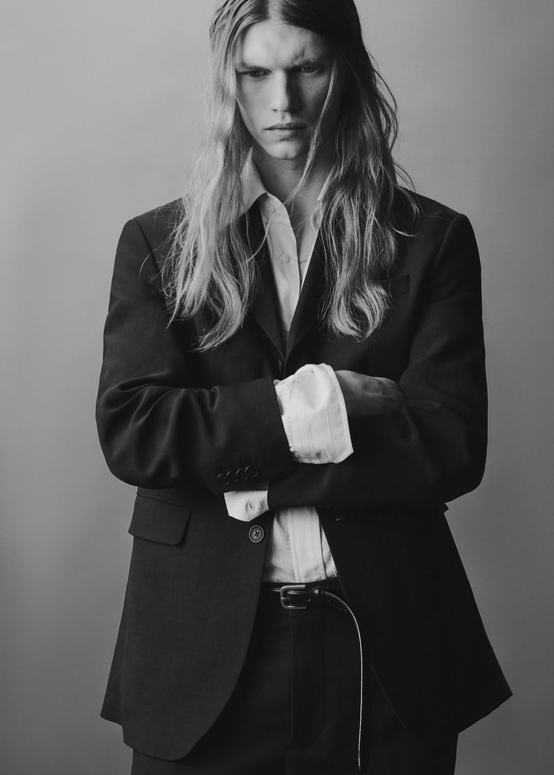 Model with long blonde hair wearing a black suit jacket and white shirt with arms crossed against a plain studio background.