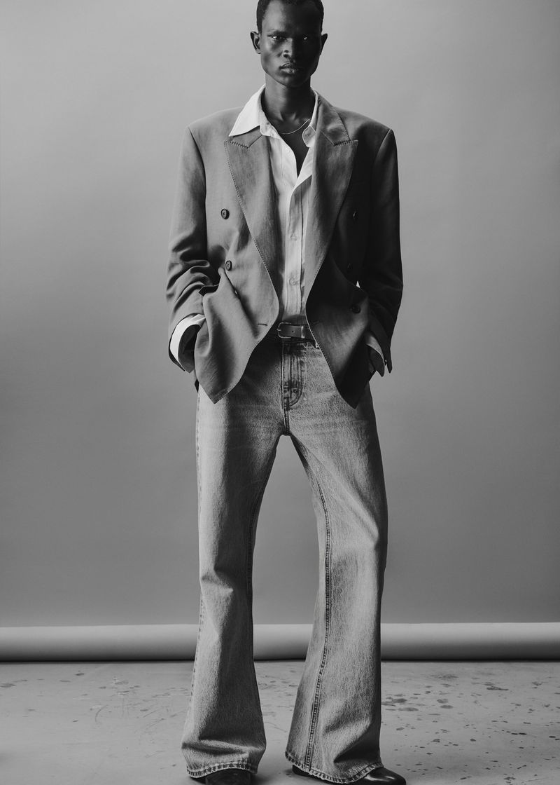 Monochrome studio shot of a Black male fashion model in a blazer, white shirt, and light blue jeans, hands in pockets, full length.
